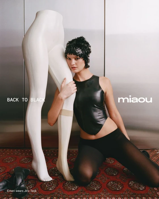 Model kneeling beside a white mannequin leg wearing a black top, tights, and a sequined cap on a patterned rug.