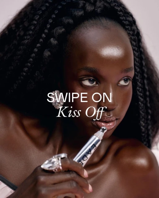 Close-up beauty shot of a woman with braids applying lip product, showcasing silver packaging, and ring jewelry against a light background.