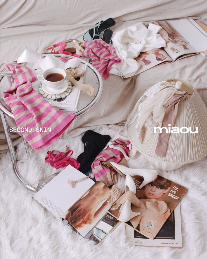 Still life on a bed with women's clothing, shoes, jewelry, magazines, a cup of tea, and a lamp creates a relaxed, fashion-themed scene.