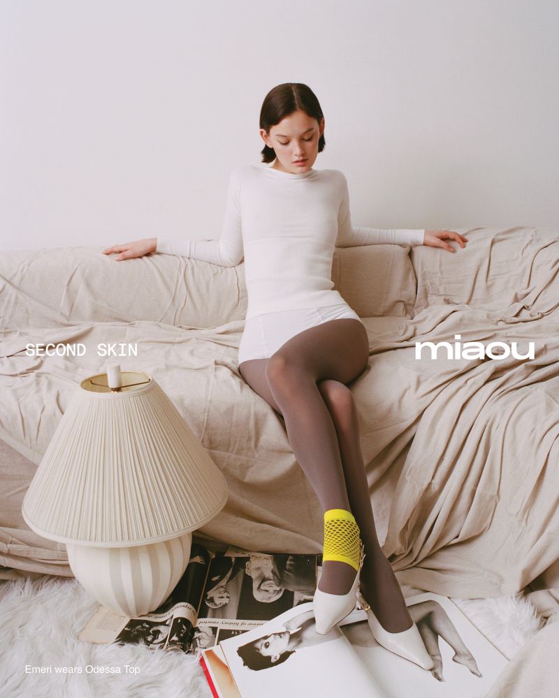 A woman poses on a tan couch in a white long-sleeve shirt, white shorts, sheer grey tights, yellow socks, and white heels with magazines nearby.