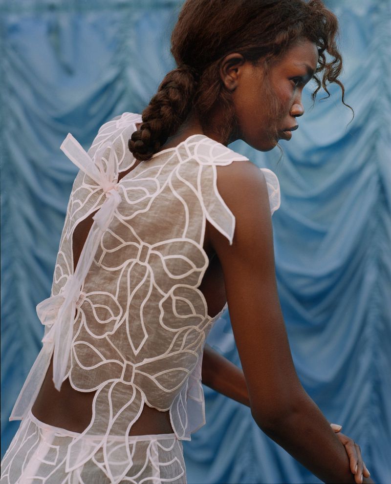 A black female model in a white lace top with a bow design poses against a blue draped fabric backdrop in a fashion editorial.