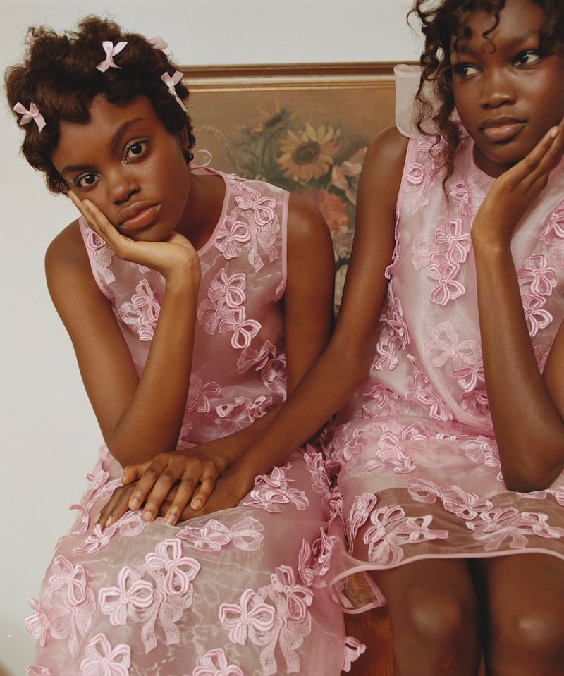 Two young Black women pose indoors wearing matching pink dresses with bows, complemented by hair accessories, against a neutral backdrop.