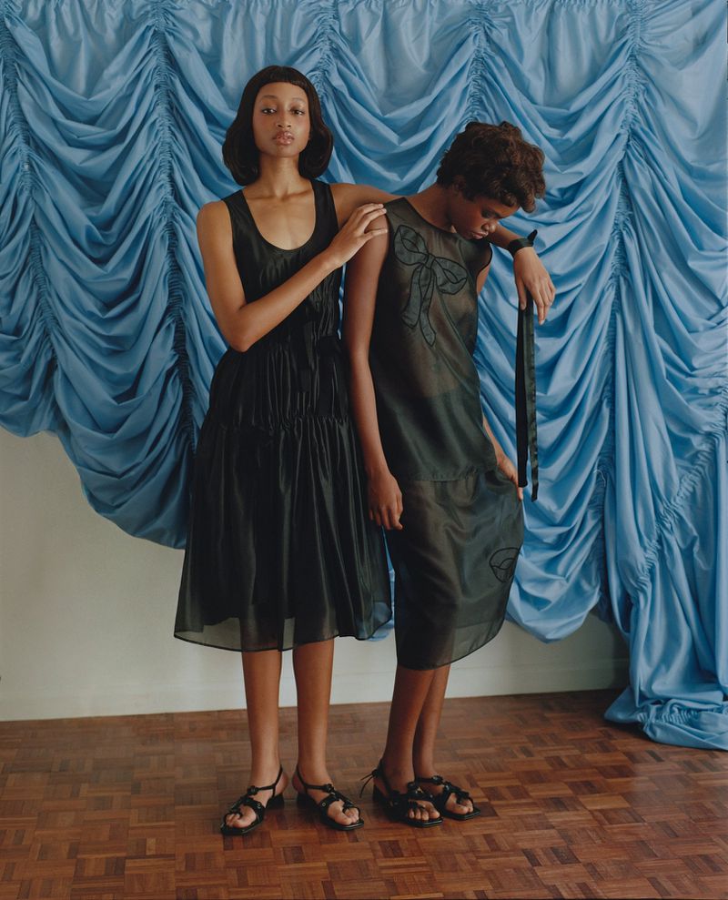 Two Black women in black dresses pose against a blue draped fabric backdrop with a wood floor in a fashion photography style studio.