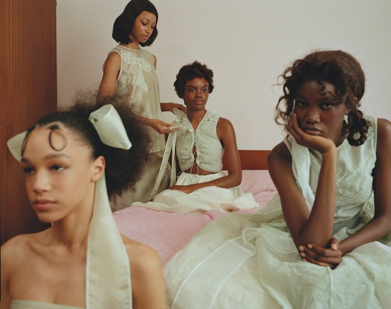 A group of four Black women wearing light green dresses are gathered indoors on a pink bed in a fashion editorial image.