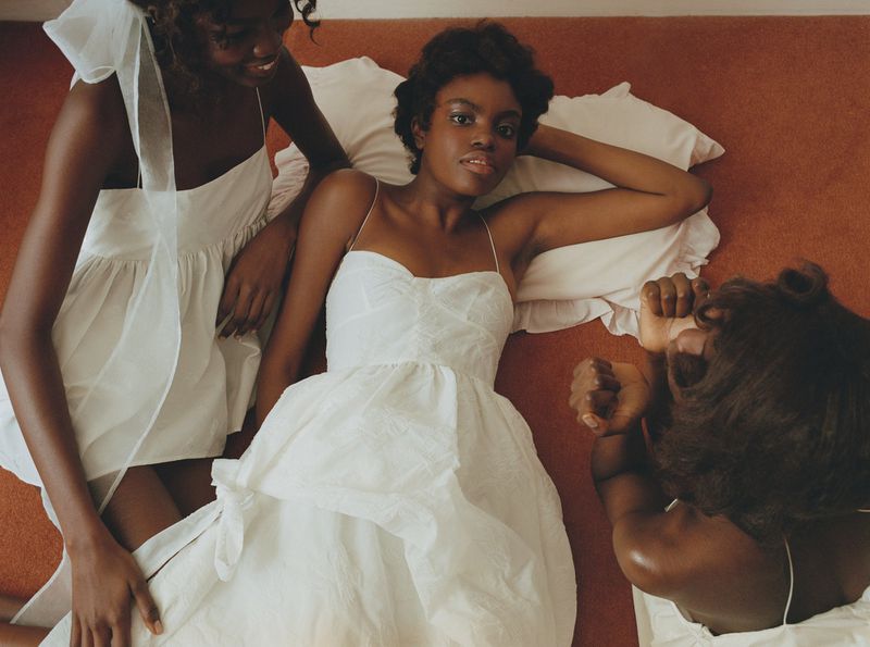 Three women in white dresses are lying on a bed with an orange carpet. One woman is looking at the camera.