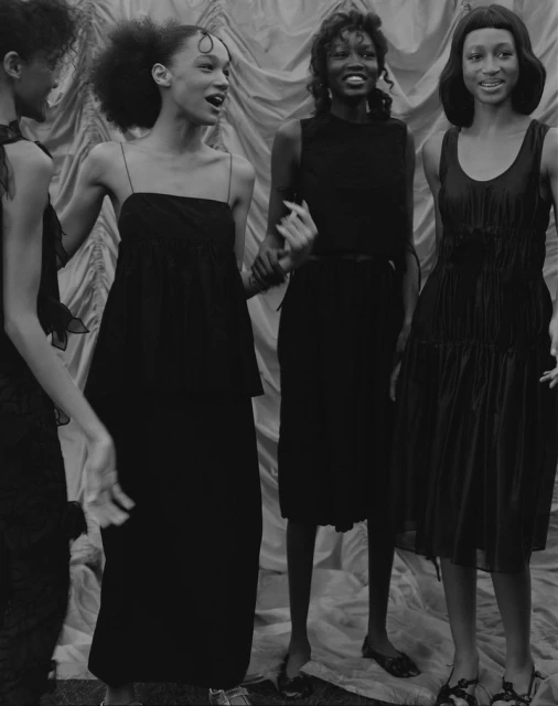 Four Black women in black dresses stand in front of a draped backdrop, monochrome fashion shot.