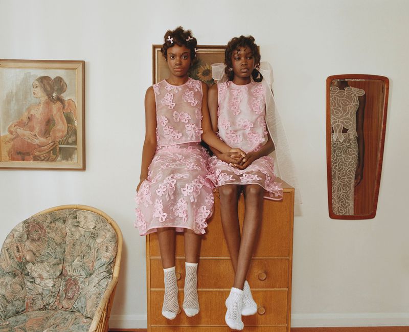 Two young Black girls in matching pink dresses sit on a wooden dresser in an interior setting, with vintage paintings and a chair visible.
