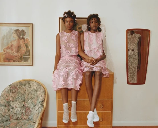 Two young Black girls in matching pink dresses sit on a wooden dresser in an interior setting, with vintage paintings and a chair visible.