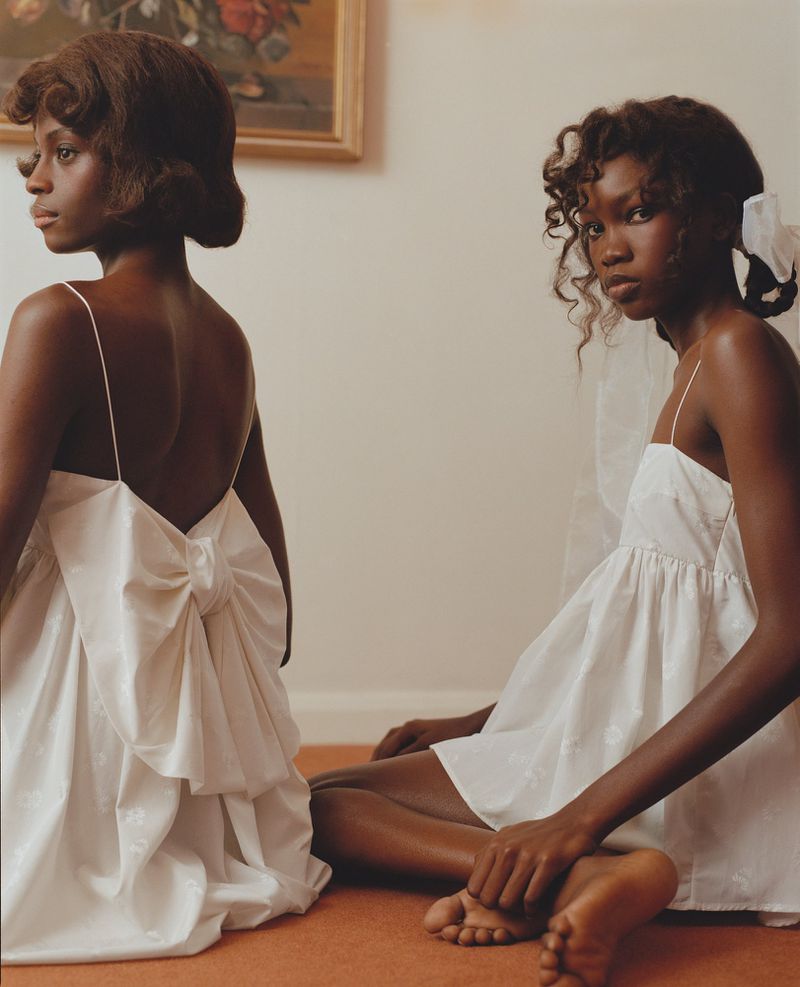 Two African American women in white dresses sit on an orange surface in a brightly lit indoor shot. One dress has a large bow.