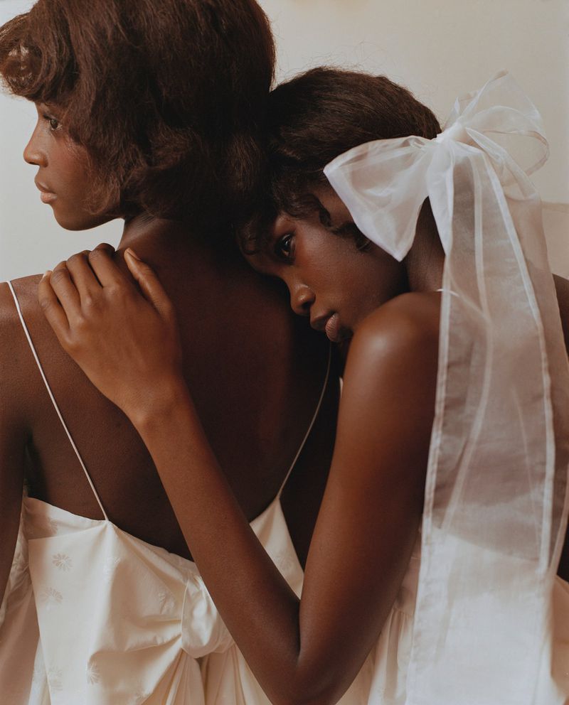 Two African American women in white dresses embrace indoors. One woman wears a sheer white bow. Soft lighting, neutral background.