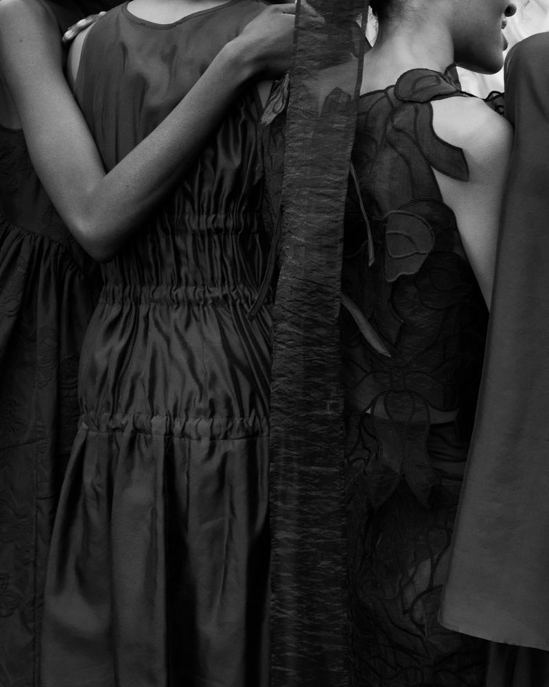 Black-and-white fashion image features a group of women in dresses, with focus on their torsos, arms, and dresses in a studio-like setting.