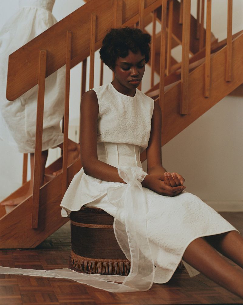 Woman sits on a fringed stool indoors, wearing an elegant white dress with a sheer sash. Stairs and another woman are visible.