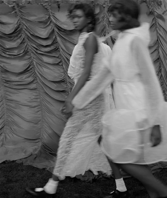 Two black women in white dresses walk outdoors, showcasing fashion in a black-and-white aesthetic with a draped fabric backdrop.