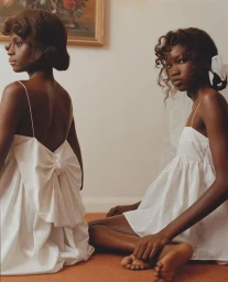 Two Black female models in white slip dresses with bows seated on a floor for Cecilie Bahnsen Pre-Fall 2026