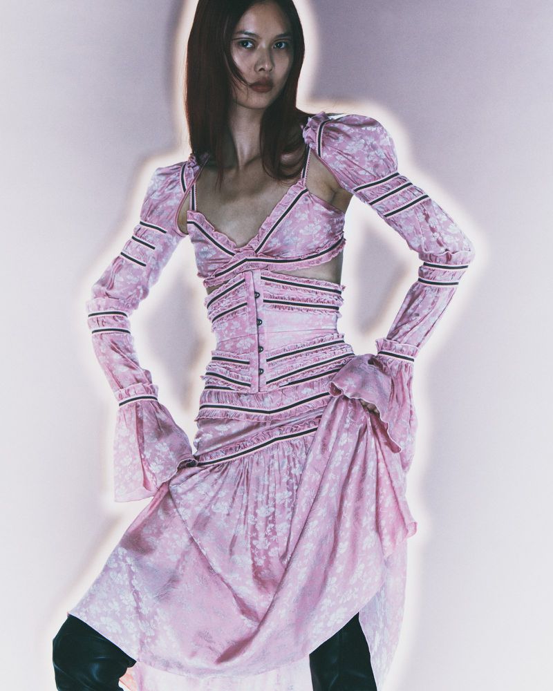 Woman posing in a pink floral-patterned dress with long sleeves and black boots, against a soft pink background, fashion photography.
