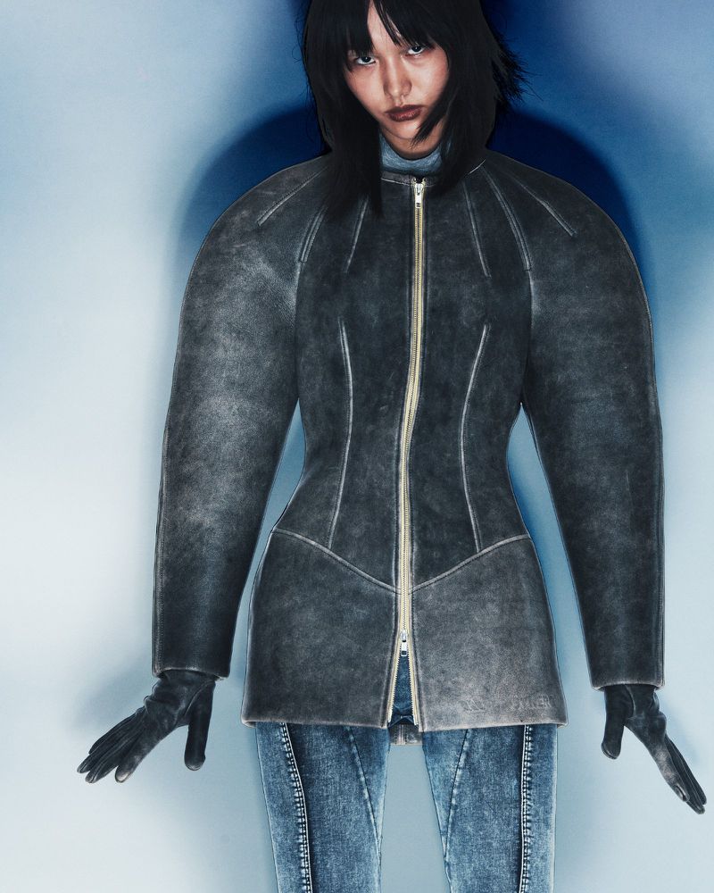 Model in dark structured zip-up jacket and blue jeans standing against a blue gradient studio background.