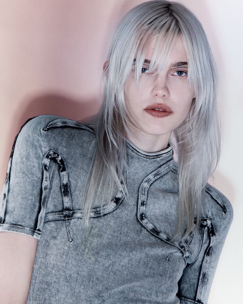 A young adult woman with long, silver hair poses indoors, wearing a structured gray top against a soft pink and white background.