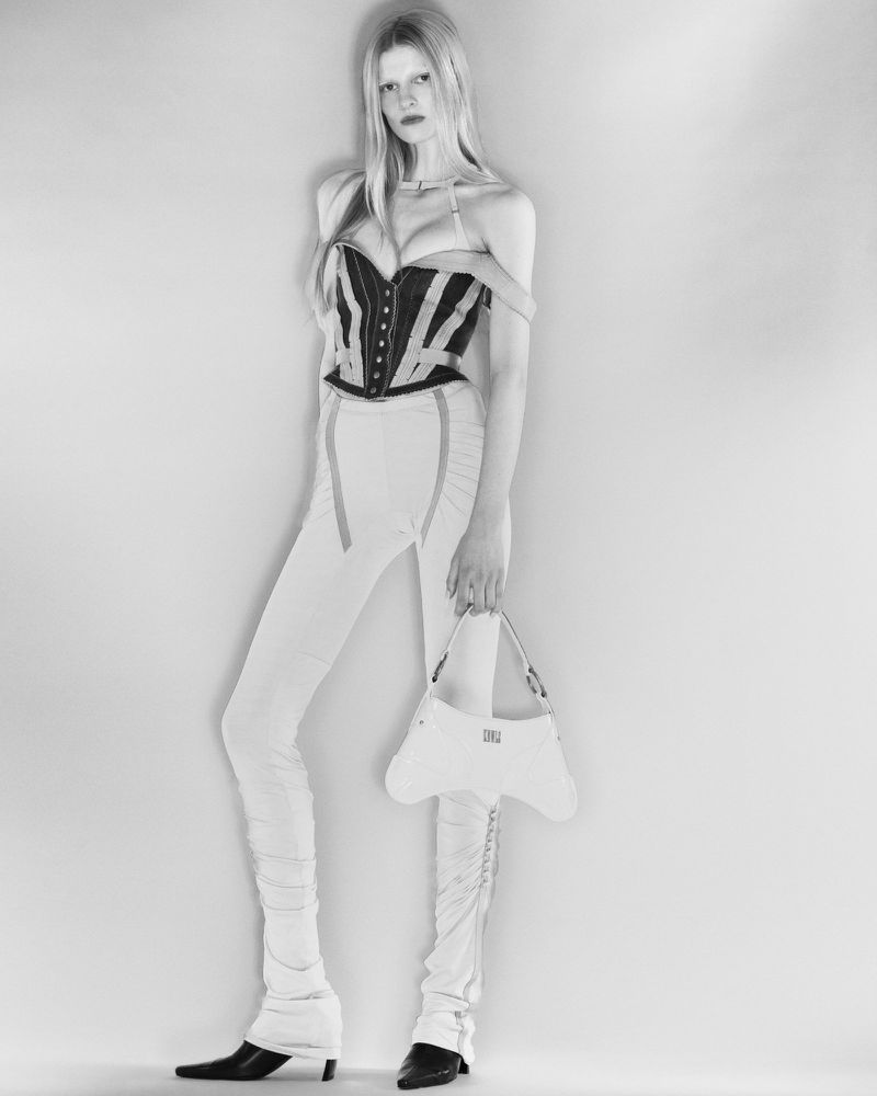 A woman poses in a corset top and white pants, holding a white bag, in a studio shot against a white backdrop, in a fashion editorial.