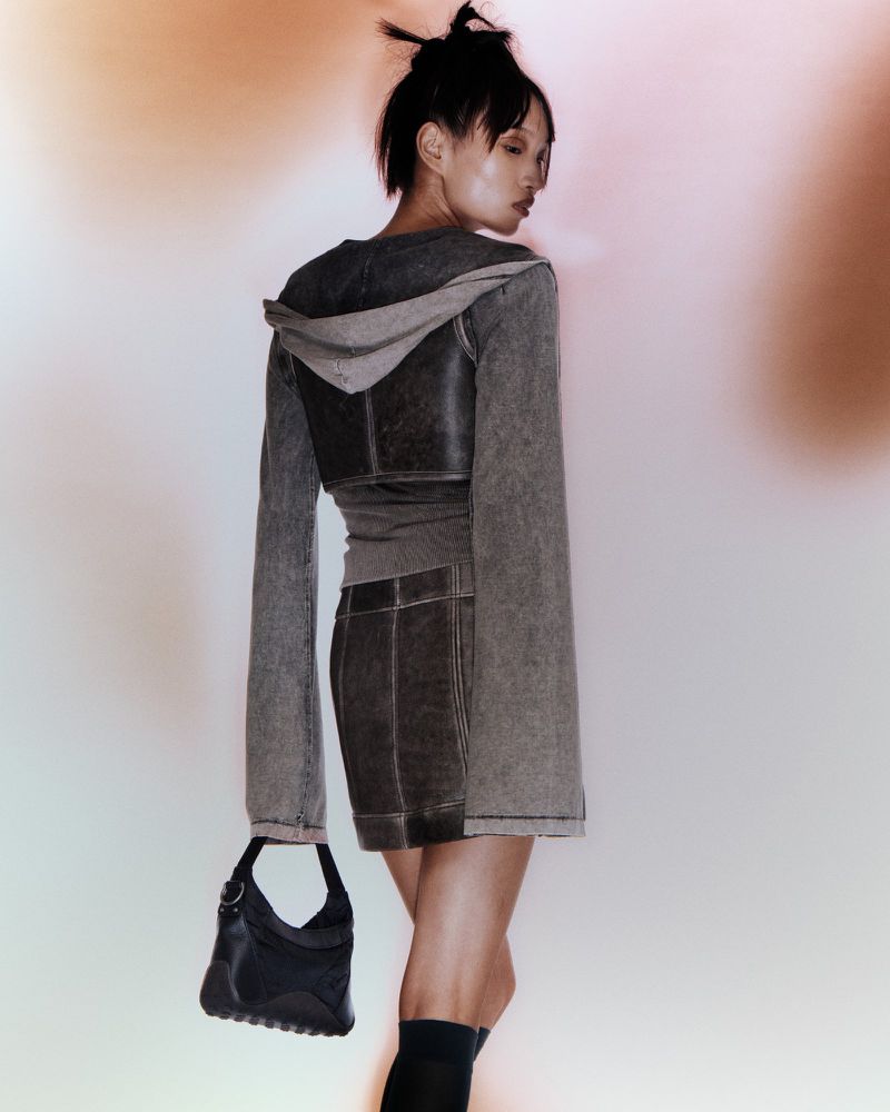 Back view of an Asian woman posing in a grey hooded top and skirt with a black purse against a soft, diffused background.