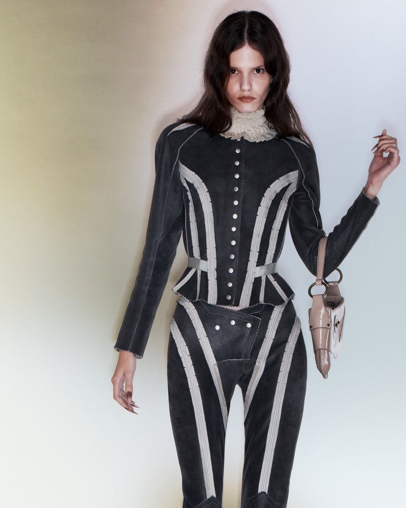 A slender woman in a striped jacket and pants poses in a studio with a handbag, showcasing a high-fashion editorial style.