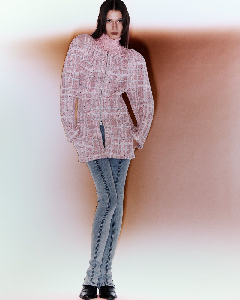 Full-length shot of a young woman posing in a pink knit jacket with light wash jeans in a studio with soft, diffused lighting.