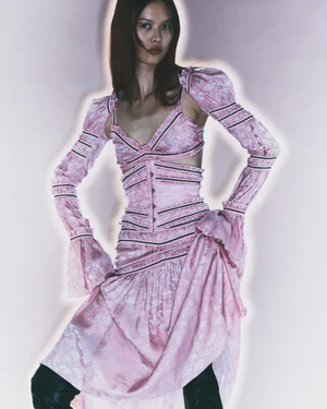Model wearing a pink patterned dress with detached long sleeves and black boots against a pale glowing backdrop.