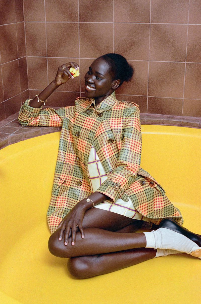 Smiling African woman in patterned coat sits in a yellow bathtub holding a yellow rubber duck.  Indoor bathroom setting, dark brown tiles.