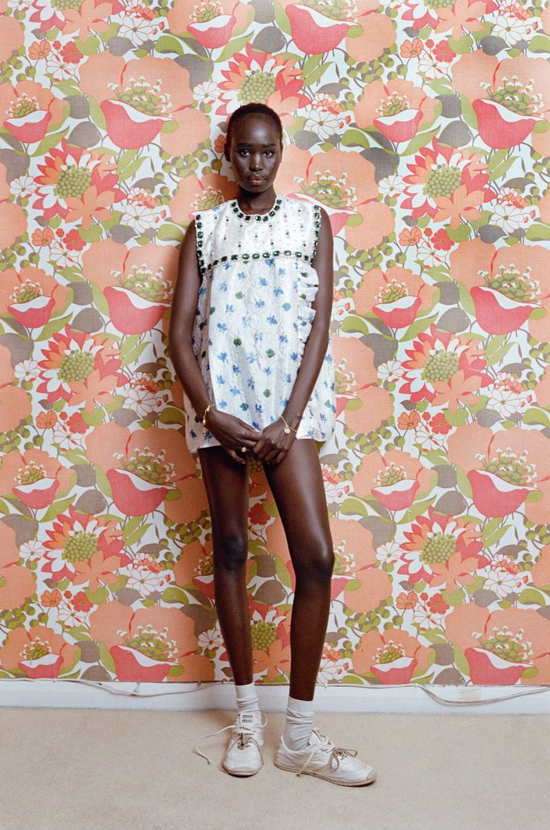 Adult female fashion model posing in a white patterned dress with vintage floral wallpaper. Full shot, eye-level angle, natural lighting indoors.