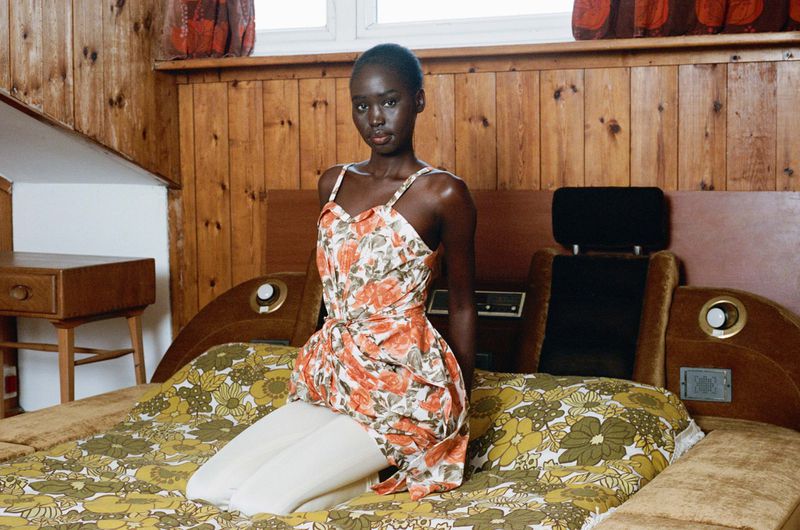 An adult female model poses kneeling on a floral-patterned bed with a wood-paneled wall as a backdrop in this brightly lit studio shot.