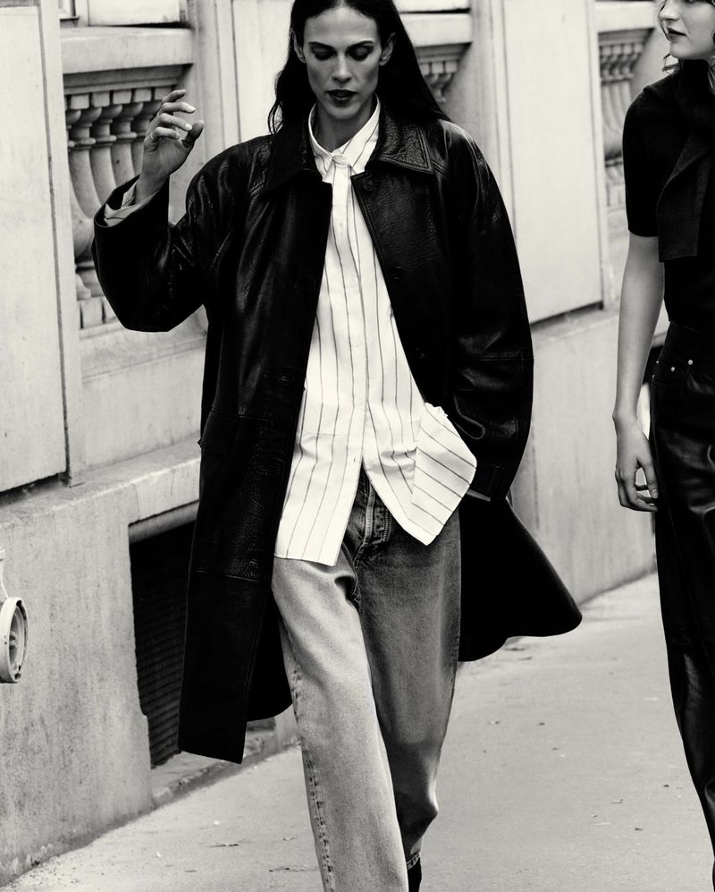 A woman walks down a city street wearing a black leather coat, light-striped shirt, and light-wash denim jeans with another woman behind.