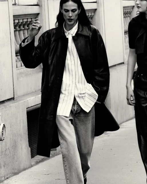 A woman walks down a city street wearing a black leather coat, light-striped shirt, and light-wash denim jeans with another woman behind.