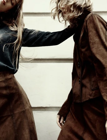 Two women in retro brown suede skirt suits stand close, showcasing 70s-inspired fashion against a neutral background.
