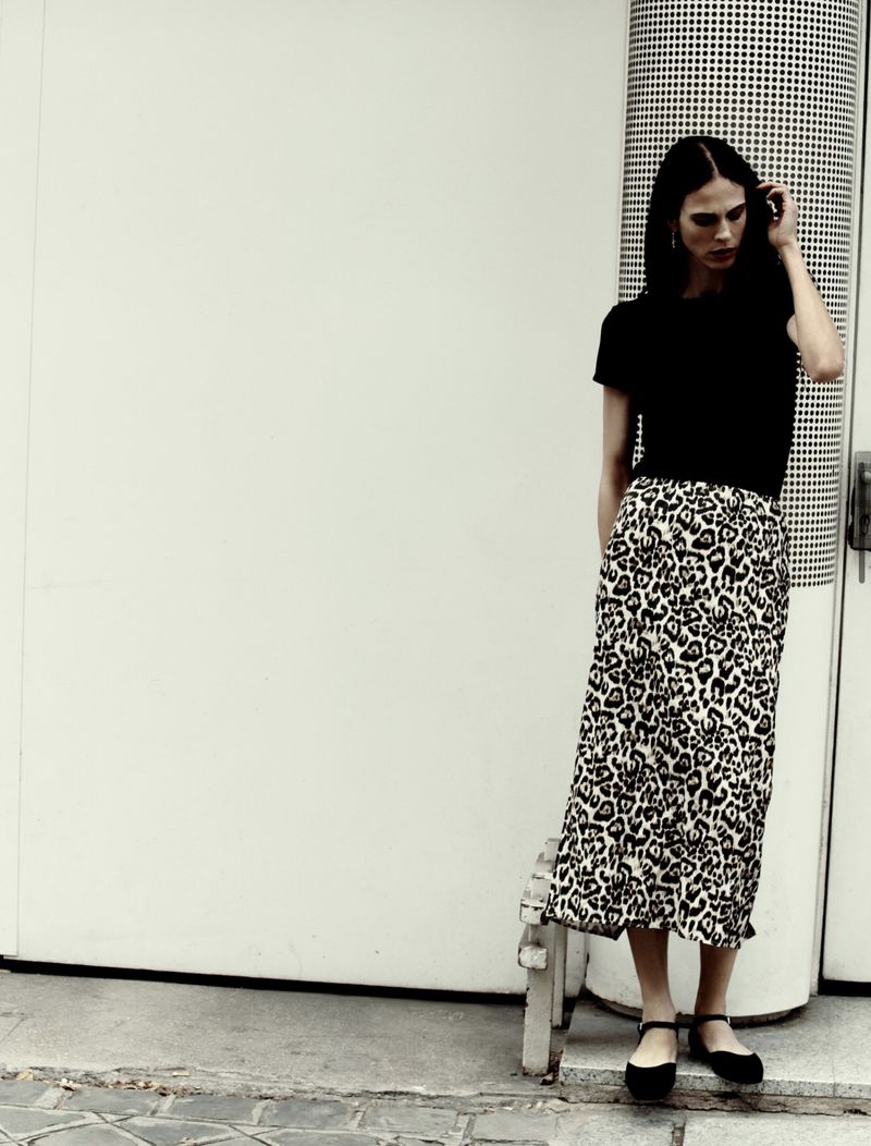 A woman stands near a wall, wearing a black top and a leopard print skirt. She is seen from the medium shot in an outdoor setting.