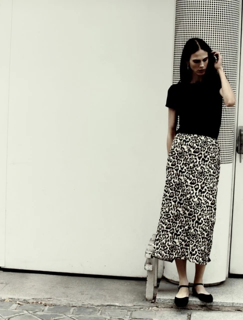 A woman stands near a wall, wearing a black top and a leopard print skirt. She is seen from the medium shot in an outdoor setting.