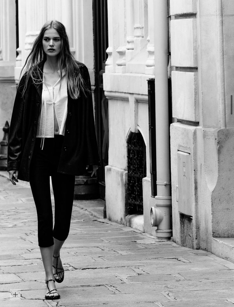 A fair-skinned woman with long hair walks on a cobblestone street wearing a black jacket, white top, and sandals in an urban setting.