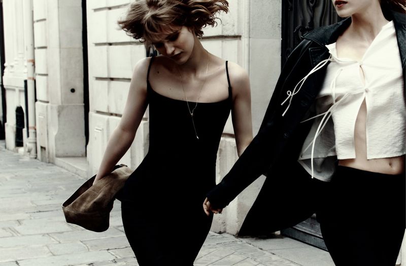 Two women walk on a cobblestone street. One in a black dress carries a jacket; the other wears a white blouse and black jacket.