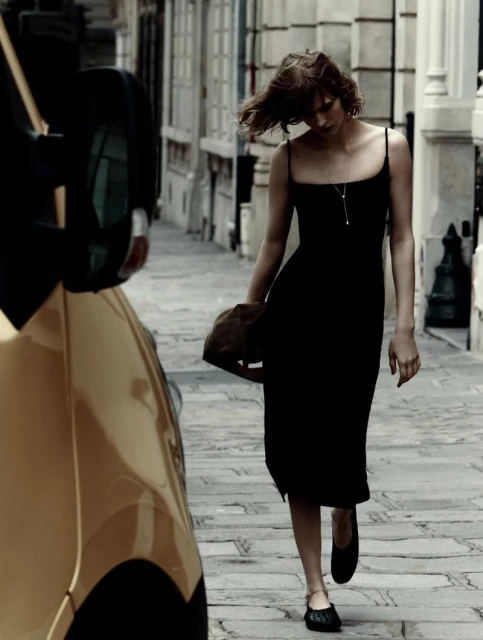 Woman in black dress walks along cobblestone street with yellow vehicle; fashion editorial conveys urban style and movement.