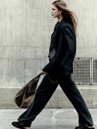 Model walking outdoors wearing dark blazer and trousers, carrying a large brown suede bag against a concrete building wall.