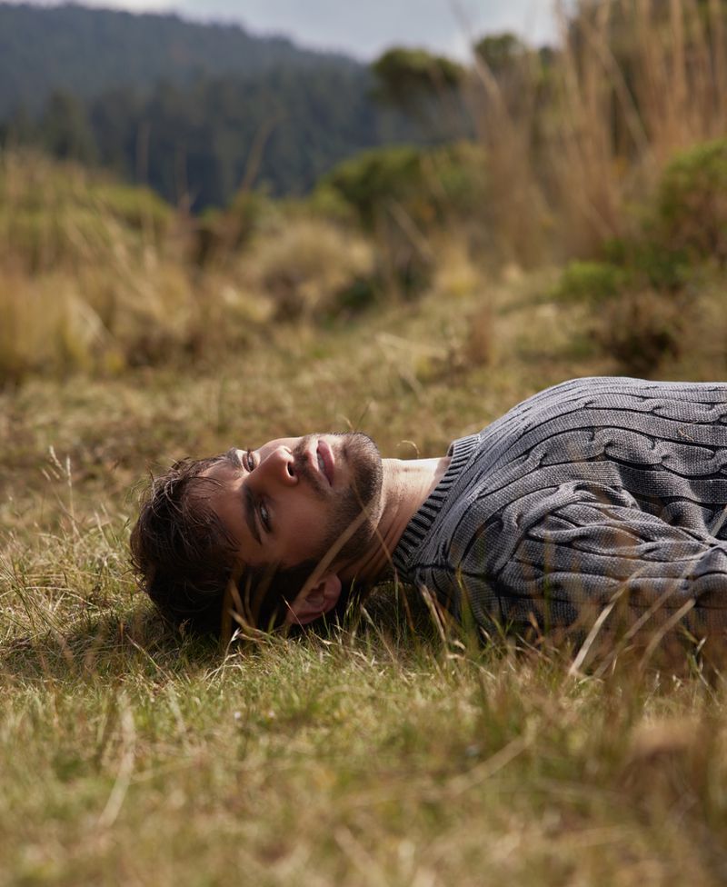 A man with dark hair wearing a grey sweater lies in the grass in a rural outdoor landscape with a forested area in the background.