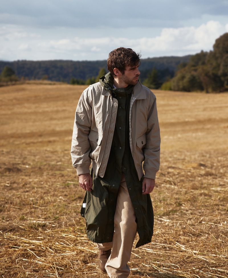 An adult male is walking across a field of dry grass wearing a khaki jacket, a long green coat, and beige pants in natural light.