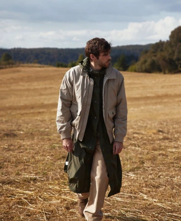 An adult male is walking across a field of dry grass wearing a khaki jacket, a long green coat, and beige pants in natural light.