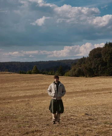 A man wearing a grey bomber jacket and dark trousers standing in a wide dry field under a dramatic cloudy sky.