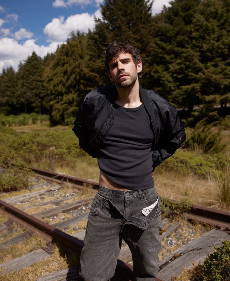 Adult male fashion portrait on train tracks in forest: man in black jacket, t-shirt, and grey jeans poses in outdoor setting.