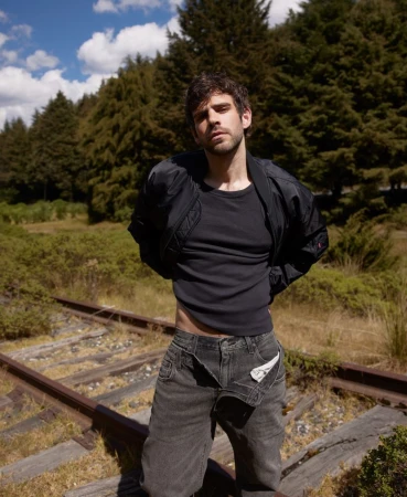 Adult male fashion portrait on train tracks in forest: man in black jacket, t-shirt, and grey jeans poses in outdoor setting.
