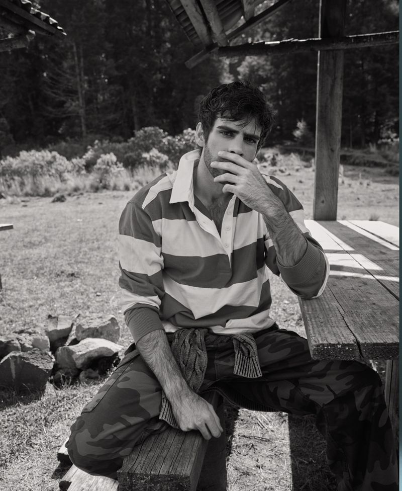 Monochrome portrait of a young adult man sitting on a wooden bench wearing a striped shirt and camouflage pants, with a forest backdrop.