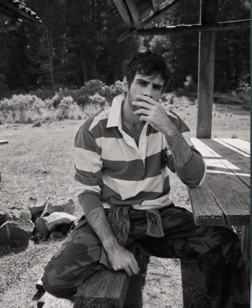 Monochrome portrait of a young adult man sitting on a wooden bench wearing a striped shirt and camouflage pants, with a forest backdrop.