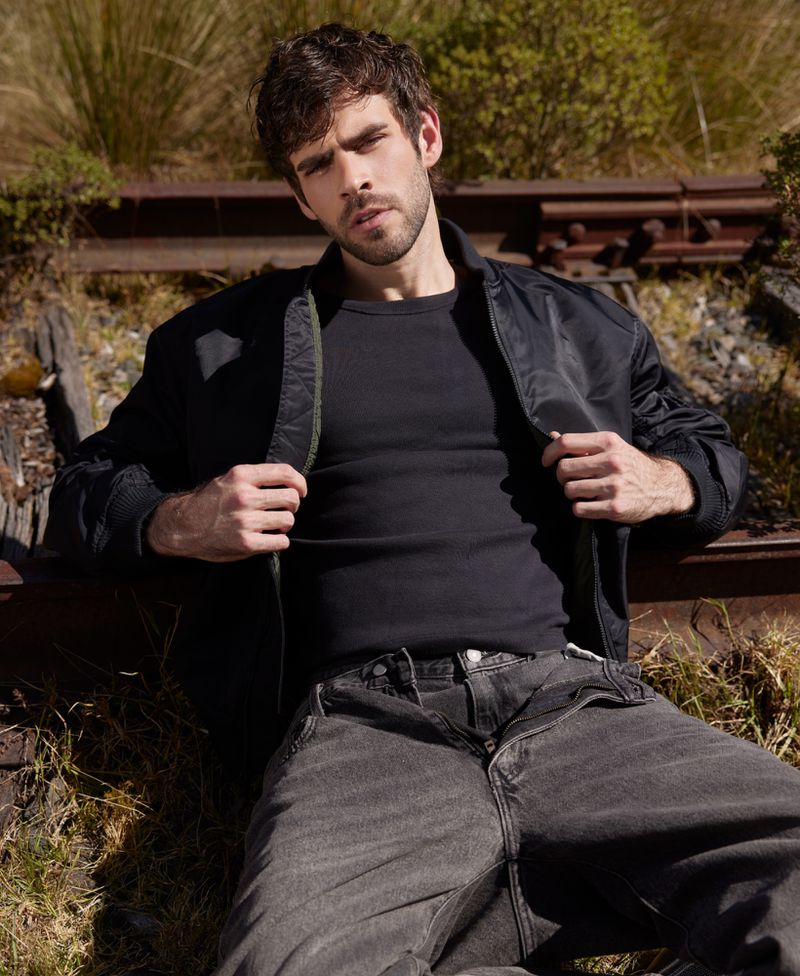 A brunette adult male in a black jacket and jeans poses on train tracks surrounded by greenery in an outdoor environment.