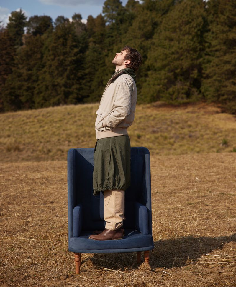 Man stands on a blue armchair in a field, looking up at the sky. Tall evergreen trees are visible in the background on a sunny day.