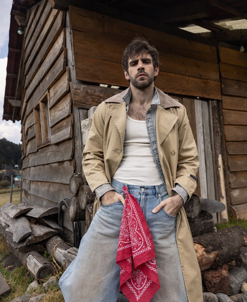 Portrait of a man in blue jeans and a trench coat stands in front of a wooden cabin, showcasing rustic outdoor fashion in daylight.