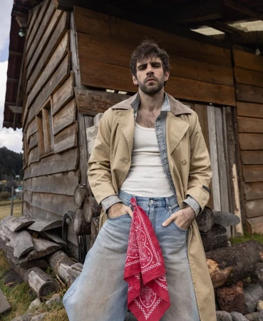 Portrait of a man in blue jeans and a trench coat stands in front of a wooden cabin, showcasing rustic outdoor fashion in daylight.